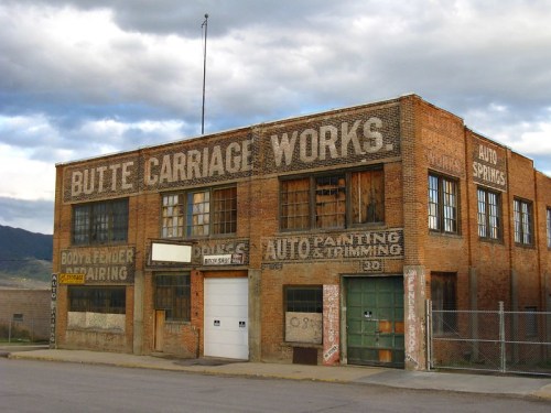 Historic brick building with faded signage for "Butte Carriage Works" and various auto services, under a cloudy sky.