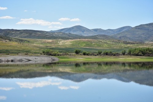 A serene landscape featuring a calm lake reflecting mountains and blue skies, surrounded by green hills and trees.
