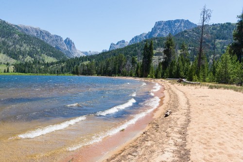 A sandy beach along a lake, with gentle waves and mountains in the background under a clear blue sky.