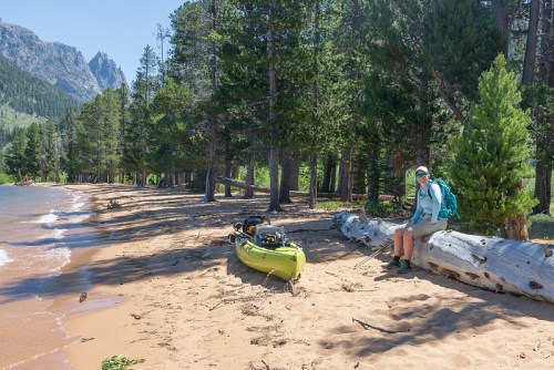 A person sits on a log by a sandy beach, with a kayak nearby and trees and mountains in the background.