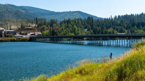 A wooden bridge spans a calm blue river, surrounded by lush green hills and trees under a clear blue sky.