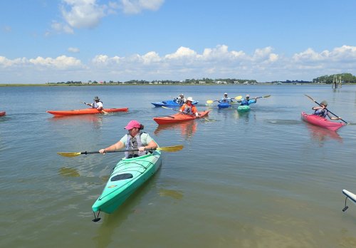 A group of people kayaking in colorful kayaks on a calm waterway under a blue sky with fluffy clouds.