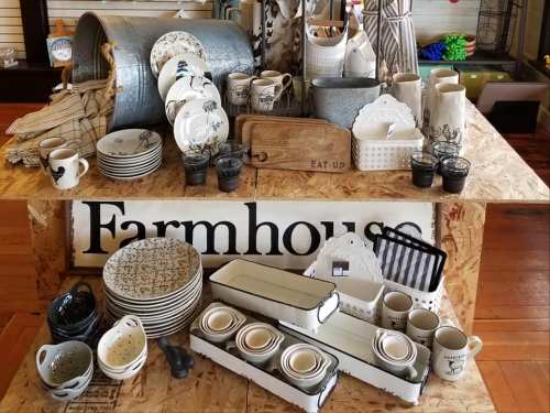 A display of various farmhouse-style kitchenware, including plates, mugs, and decorative items on a wooden table.