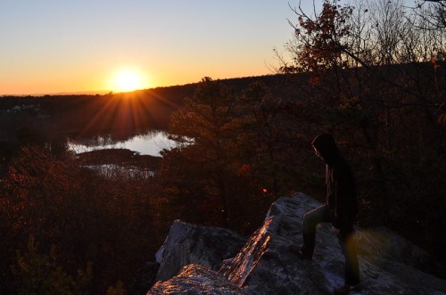 A person stands on a rock at sunset, overlooking a serene landscape with trees and a reflective body of water.