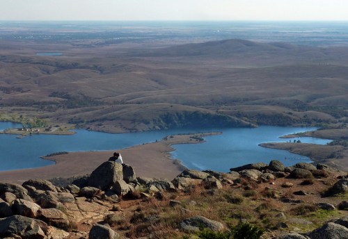 A person sits on a rock overlooking a vast landscape of hills and lakes under a clear sky.