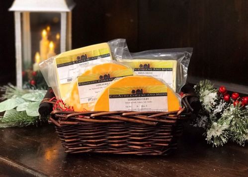 A woven basket filled with various cheese packages, decorated with festive greenery and a lantern in the background.