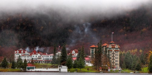 A scenic view of a historic hotel by a lake, surrounded by misty mountains and autumn foliage.