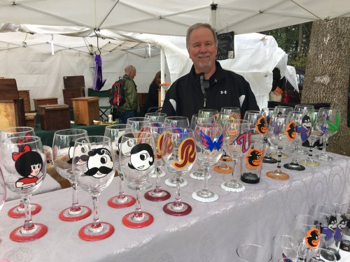A vendor smiles behind a display of hand-painted glasses featuring various cartoon characters at a market.
