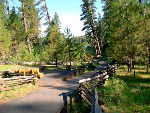 A winding path through a forest with tall trees and wooden fences, leading to a serene lake in the distance.
