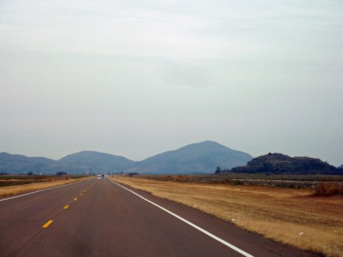 A wide, empty road stretches towards distant, hazy mountains under a cloudy sky. Brown grass lines the roadside.