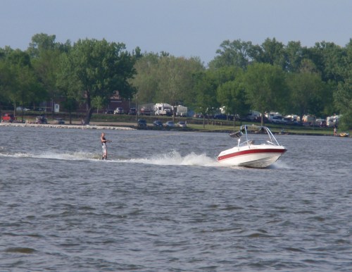 A person water skiing behind a boat on a lake, surrounded by trees and a shoreline in the background.