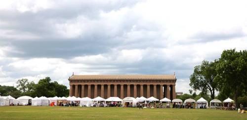 A large outdoor event with white tents set up on a grassy field, featuring a classical building in the background under cloudy skies.
