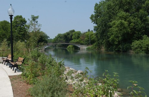 A serene river scene with a bridge in the background, surrounded by lush greenery and benches along the path.