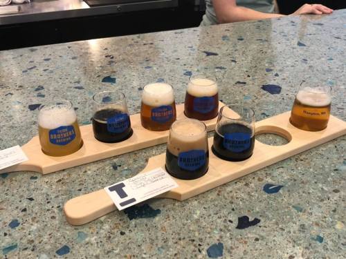 A wooden flight paddle with six small glasses of different beers on a speckled countertop.