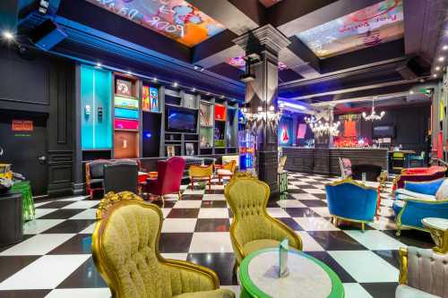 Colorful lounge with eclectic seating, a bar, and vibrant decor, featuring a checkered floor and stylish lighting.