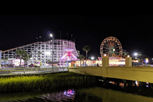 A vibrant amusement park at night featuring a roller coaster and a Ferris wheel, illuminated against a dark sky.