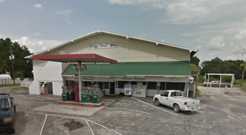 A small grocery store with a green roof, featuring a gas station and outdoor seating area. Cloudy sky in the background.