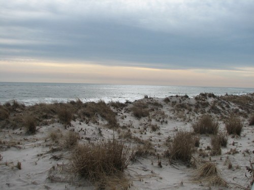 A sandy beach with sparse grass, overlooking a calm sea under a cloudy sky at dusk.