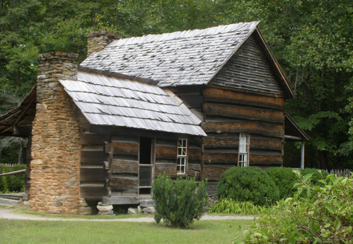 A rustic log cabin with a stone chimney, surrounded by greenery and trees.