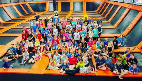 A large group of children poses together on a trampoline park, surrounded by orange and black trampolines.