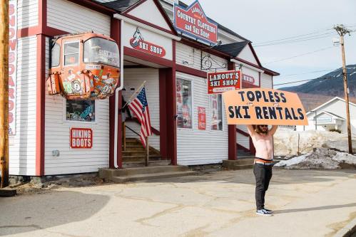 A man holds a large sign reading "Topless Ski Rentals" outside a ski shop with a gondola and American flag.