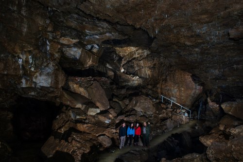 A group of people stands in a rocky cave with stalactites, surrounded by dark stone formations and a small stream.