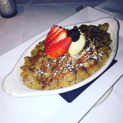 A dessert dish featuring a crumbly topping, drizzled with sauce, topped with a scoop of ice cream and fresh strawberries.