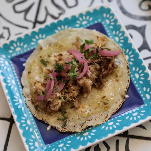 A taco on a blue plate, filled with shredded meat, topped with pink onions and cilantro, on a decorative table.