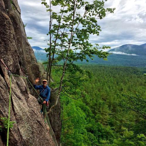 A climber waves from a rocky cliff, surrounded by lush trees and mountains under a cloudy sky.