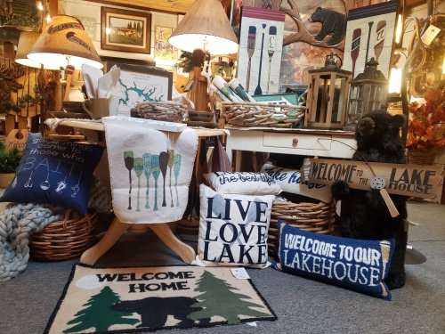 A cozy display of lake-themed decor, including pillows, signs, and tableware, in a rustic shop setting.