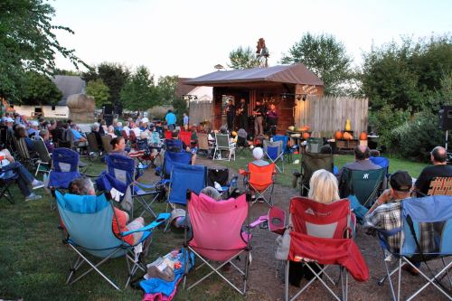 A crowd sits in folding chairs, enjoying a live performance on an outdoor stage surrounded by trees and decorations.