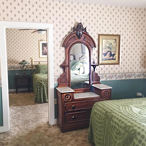 A vintage bedroom with patterned wallpaper, a wooden dresser with a mirror, and a green bedspread.
