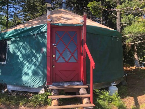 A green yurt with a red door and wooden steps, surrounded by trees in a natural setting.