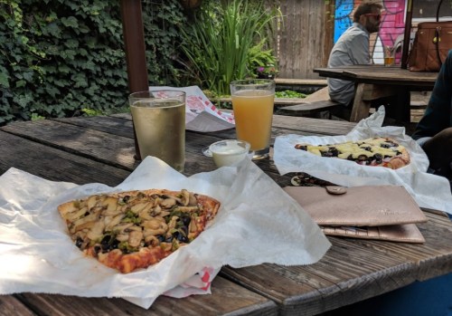 Two slices of pizza with toppings on a wooden table, accompanied by two drinks and a small container of dipping sauce.