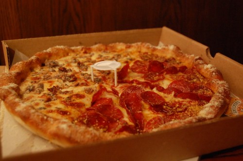 A pizza in a box, half topped with pepperoni and half with sausage, featuring a white pizza saver in the center.