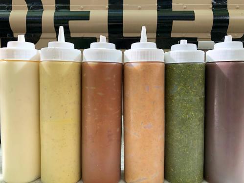 Six squeeze bottles filled with various sauces in shades of yellow, brown, orange, and green, lined up on a surface.