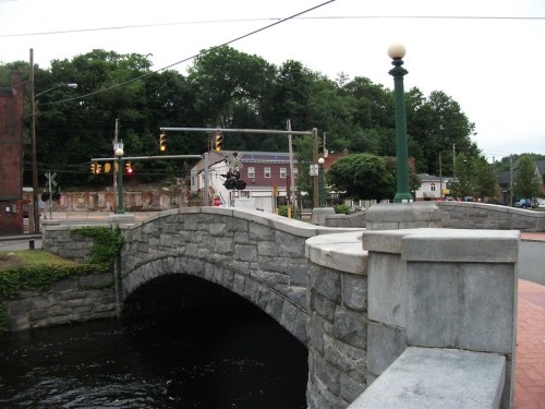 Stone bridge over a river, with traffic lights and trees in the background, under a cloudy sky.