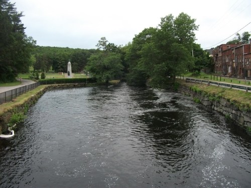 A calm river flows alongside trees and a stone wall, with a monument visible in the background under a cloudy sky.