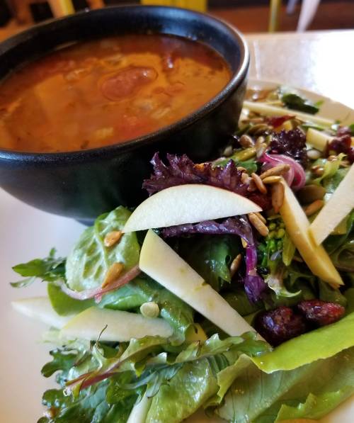 A bowl of soup next to a fresh salad topped with apple slices, seeds, and mixed greens on a white plate.