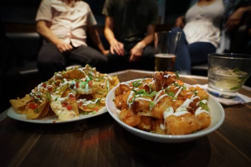 A close-up of two plates of food: nachos and crispy bites, with drinks and people blurred in the background.