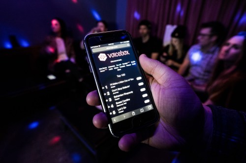 A hand holds a smartphone displaying a karaoke app, with people socializing in the background under colorful lights.