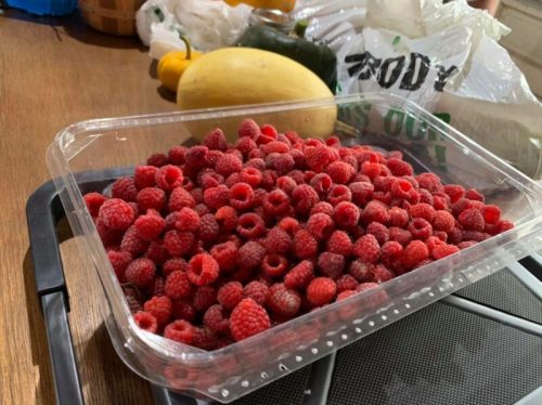 A clear container filled with fresh raspberries, with various vegetables in the background on a wooden surface.
