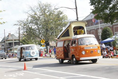 Two vintage Volkswagen vans, one orange and one blue, drive down a street with spectators and trees in the background.