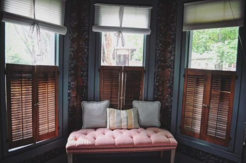 Cozy corner with a pink tufted bench, surrounded by wooden shutters and natural light from the windows.