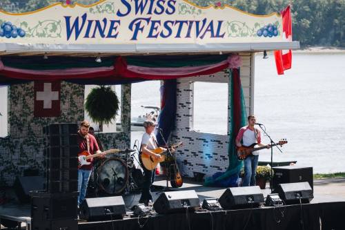 A band performs on stage at the Swiss Wine Festival, with a scenic backdrop of water and colorful decorations.