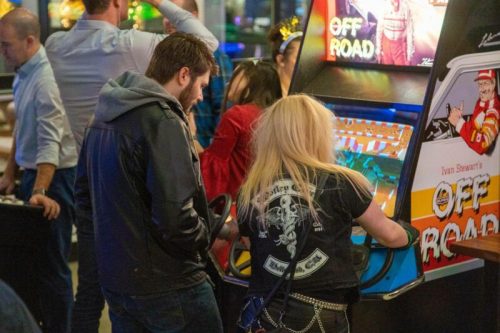 Two people play an arcade game titled "Off Road" in a lively gaming venue, surrounded by others enjoying the atmosphere.