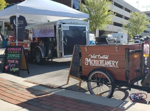 A food truck and a bicycle cart at a market, with signs for kombucha and ice cream. Trees and a parking garage in the background.