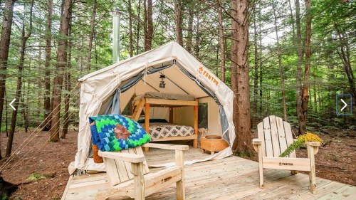 A cozy glamping tent in a forest, featuring a bed, wooden deck, and two outdoor chairs.