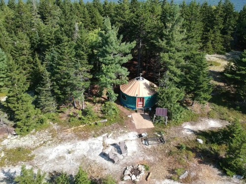 Aerial view of a yurt surrounded by trees, with a fire pit and seating area nearby on a rocky landscape.