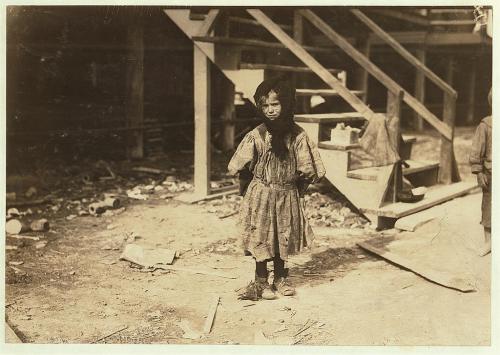 A young child stands alone in a dusty outdoor setting, wearing a striped dress and looking directly at the camera.